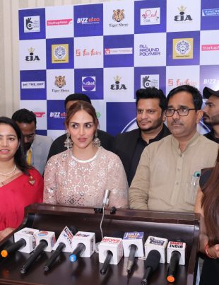 A group of people, including a woman in a white lace dress and a woman in a red saree, standing in front of a backdrop with various logos and brands, giving a press conference with multiple microphones on a podium.